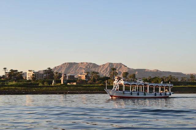 A serene view of the Nile River near Luxor, with a traditional Egyptian boat gently cruising past lush greenery. In the background, the rugged cliffs of the Theban hills hint at the ancient necropolises hidden within, including the famous Valley of the Kings.