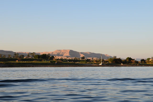 A tranquil scene on the Nile River at sunset, capturing the golden-hued Theban hills in the distance. The lush riverbank contrasts with the arid cliffs, marking the gateway to ancient wonders such as the Valley of the Kings and the temples of Luxor's west bank.