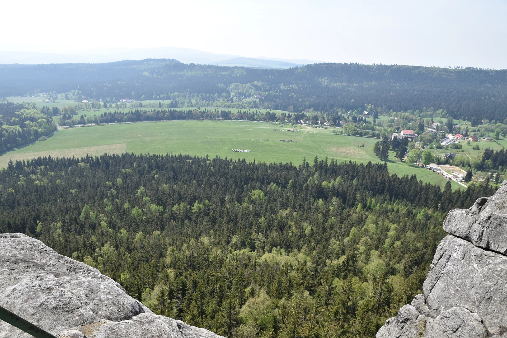 A breathtaking vantage point from the Table Mountains near Kudowa Zdroj showcases a patchwork of dense forests and vast green plains below. The landscape unfolds with scattered rural settlements nestled among the natural beauty.