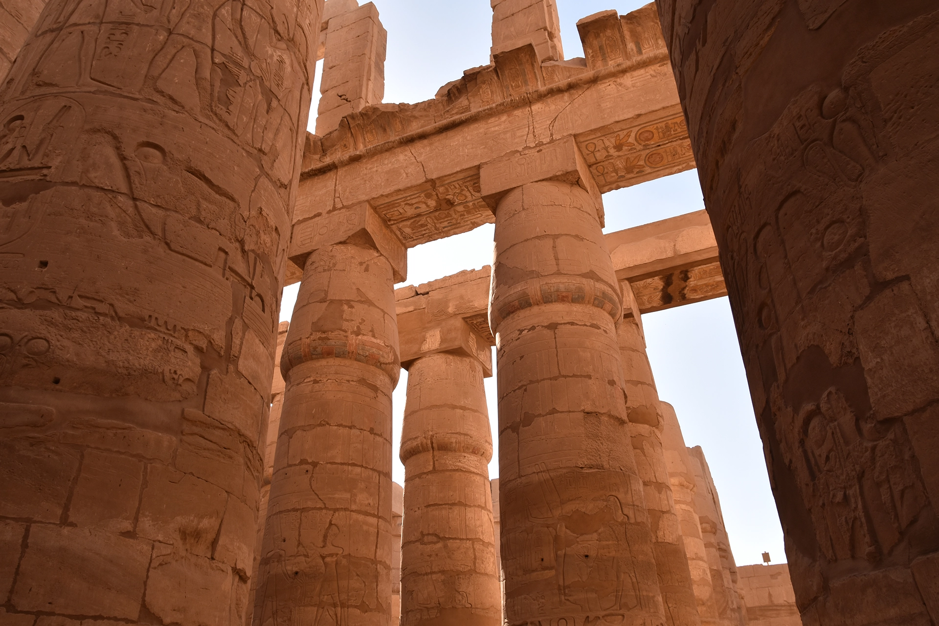 A captivating scene within Karnak Temple's hypostyle hall, where intricately inscribed columns soar skyward, supporting ancient beams etched with hieroglyphs. Sunlight filters through the gaps, illuminating this grand testament to Egypt's architectural prowess.