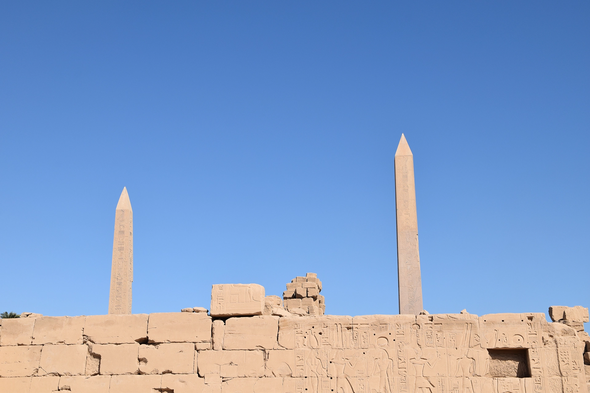 Two towering obelisks pierce the clear blue sky at the Karnak Temple complex, standing as monumental tributes to ancient Egyptian pharaohs. These intricately inscribed pillars symbolize power and divine connection, casting a legacy of awe and reverence across millennia.