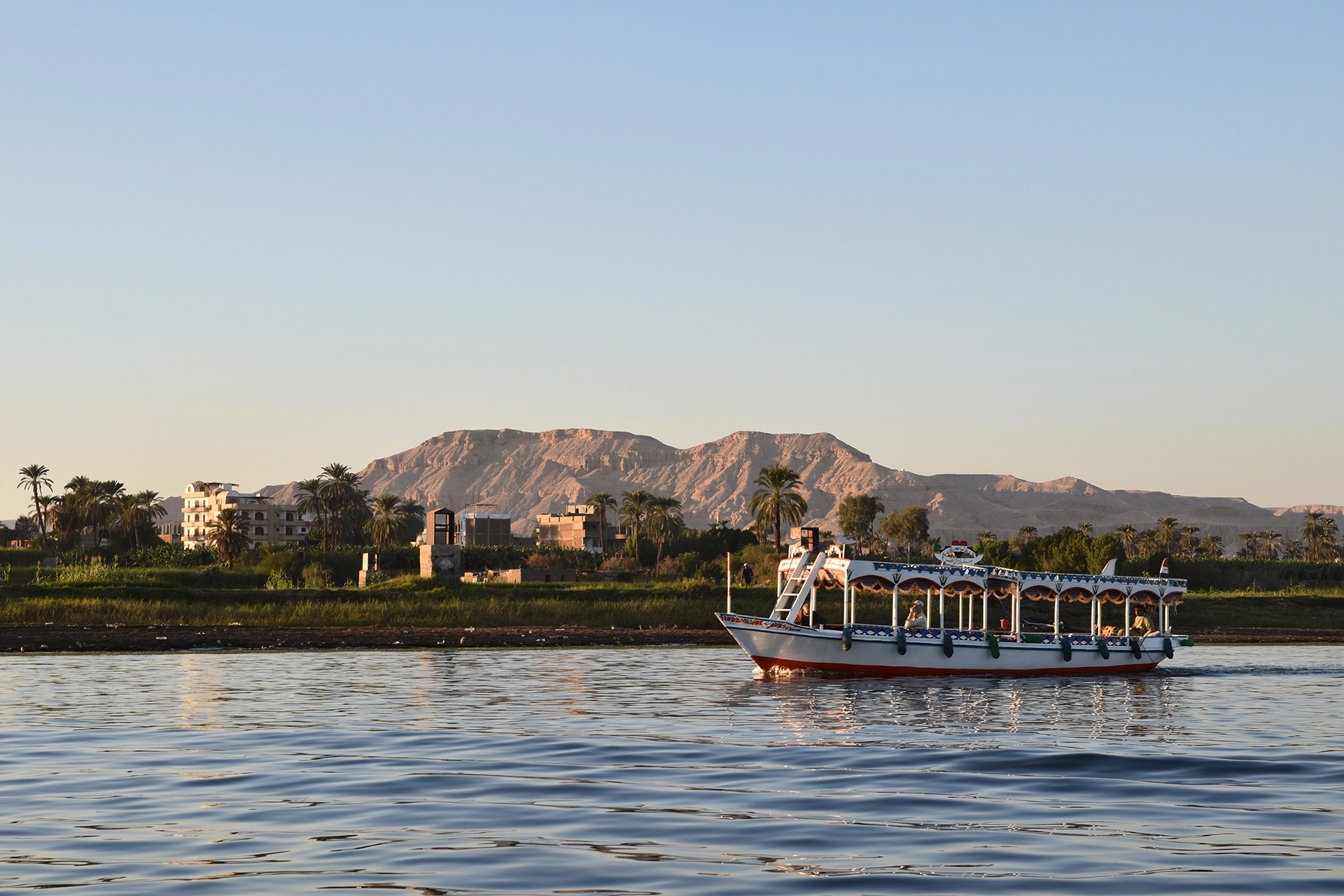 A serene view of the Nile River near Luxor, with a traditional Egyptian boat gently cruising past lush greenery. In the background, the rugged cliffs of the Theban hills hint at the ancient necropolises hidden within, including the famous Valley of the Kings.