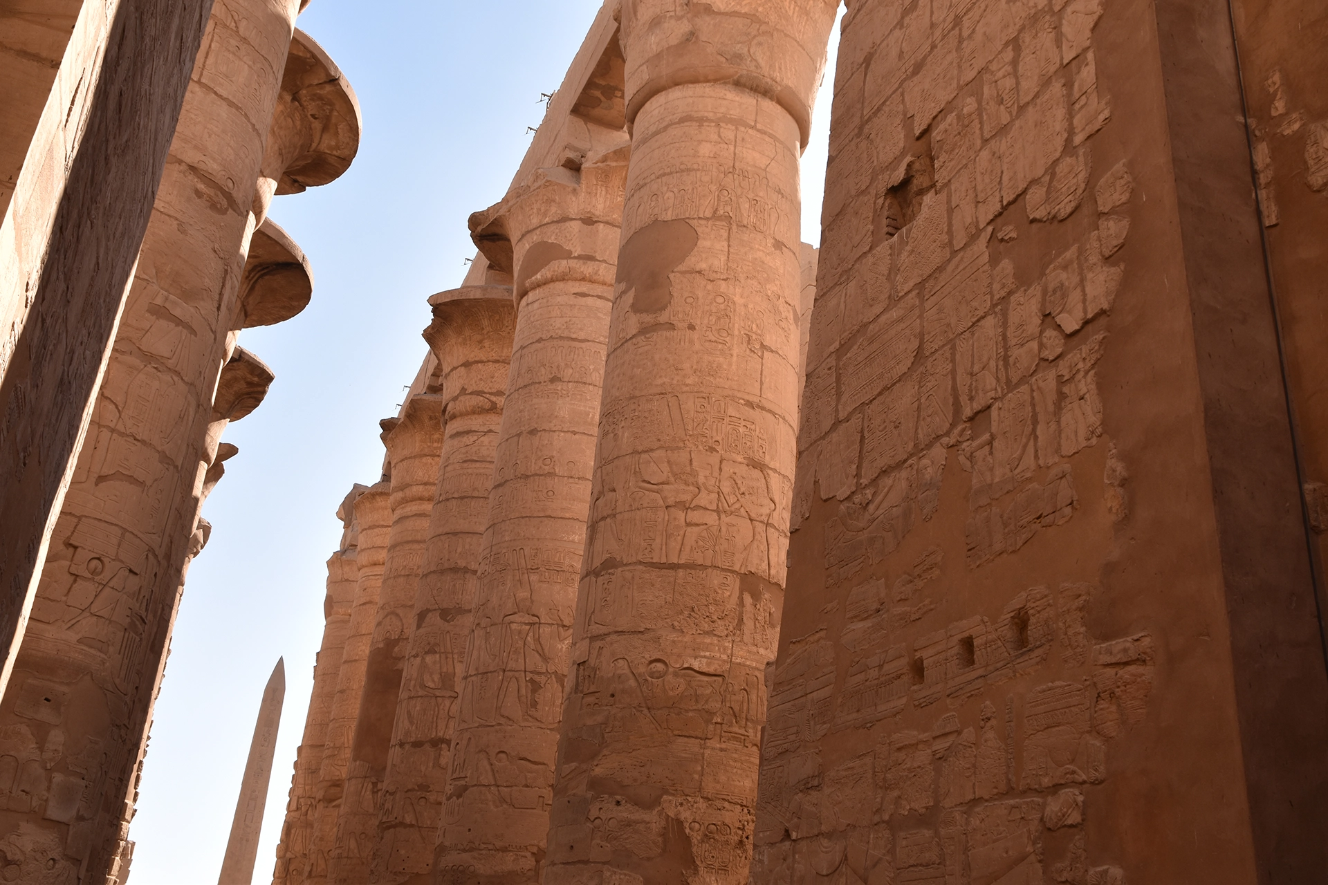 A stunning view of Karnak Temple's hypostyle hall, where colossal, hieroglyph-covered columns stretch towards the sky, framed by the distant silhouette of a timeless obelisk.