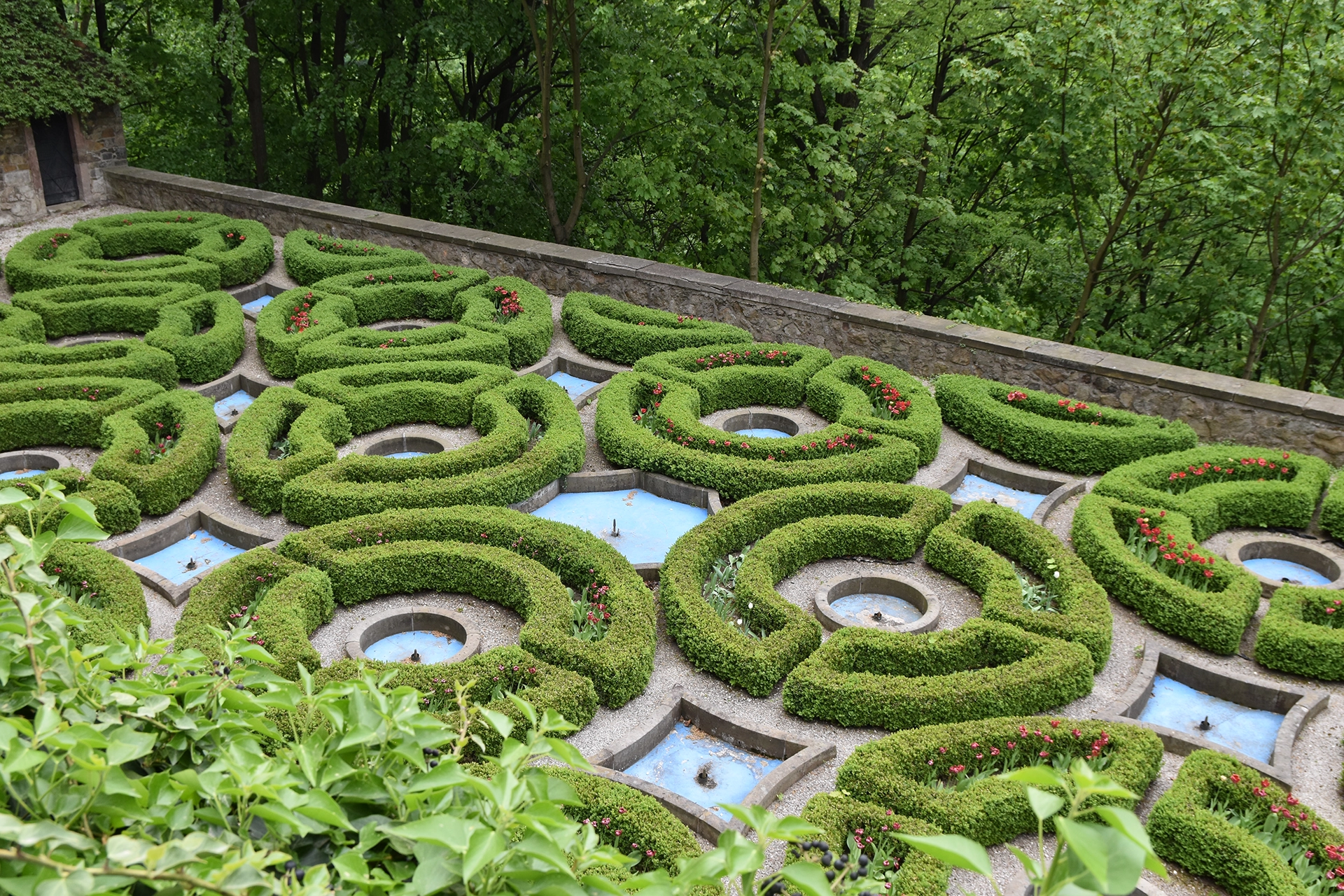 The castle's exterior gardens feature intricately designed hedges arranged in a symmetrical pattern, complemented by small fountains and vibrant flowers. This elegant landscape showcases the artistry and meticulous planning of the castle's grounds.