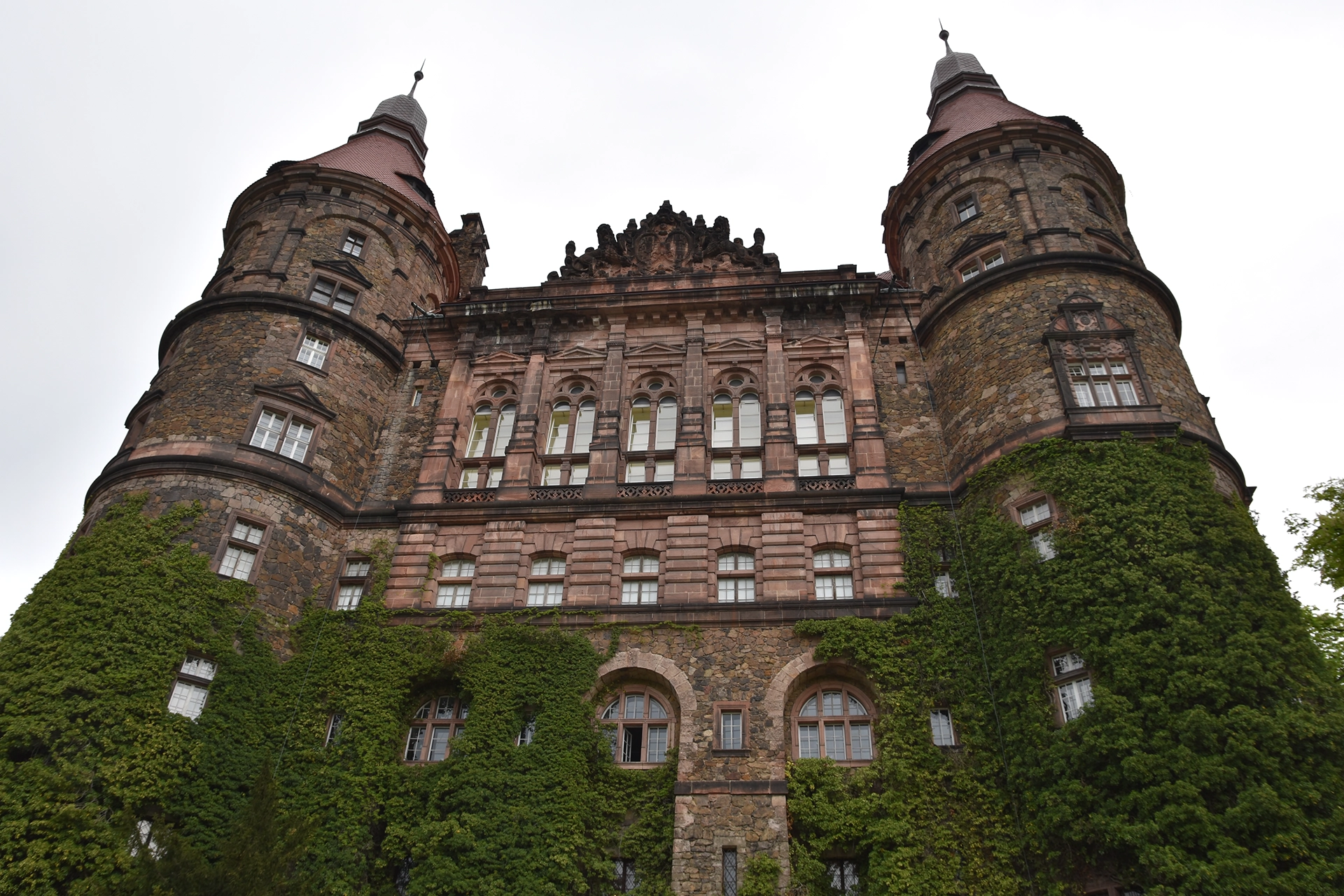 An imposing view of the castle reveals its grand facade, flanked by two cylindrical towers and adorned with lush ivy climbing the stone walls. The architecture blends medieval fortification with refined elegance, creating a captivating sight.