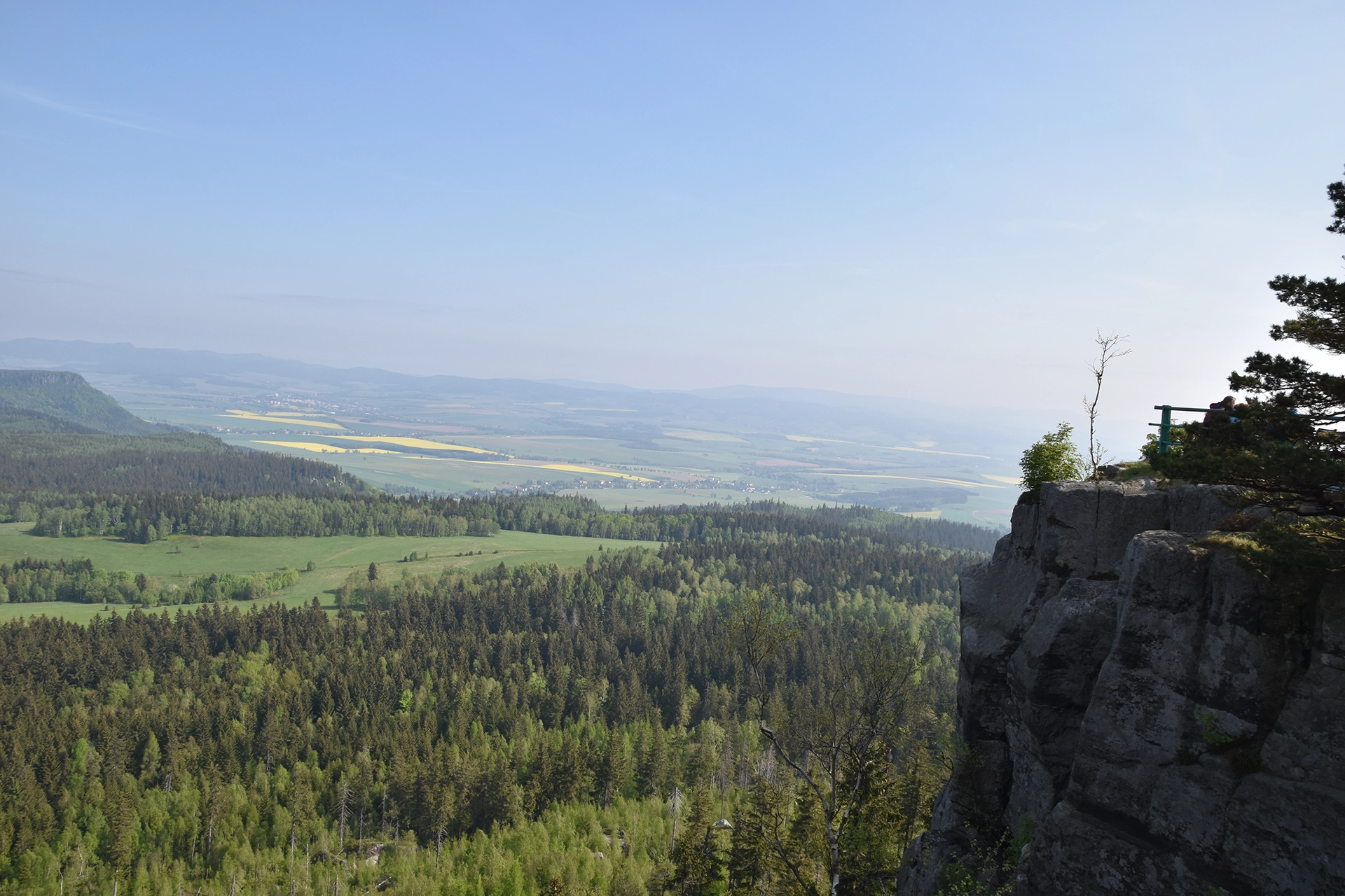 A panoramic view from the Table Mountains near Kudowa Zdroj showcases a dramatic cliff edge overlooking vast fields and forests below. The breathtaking landscape highlights the region's unique geological formations and natural beauty.