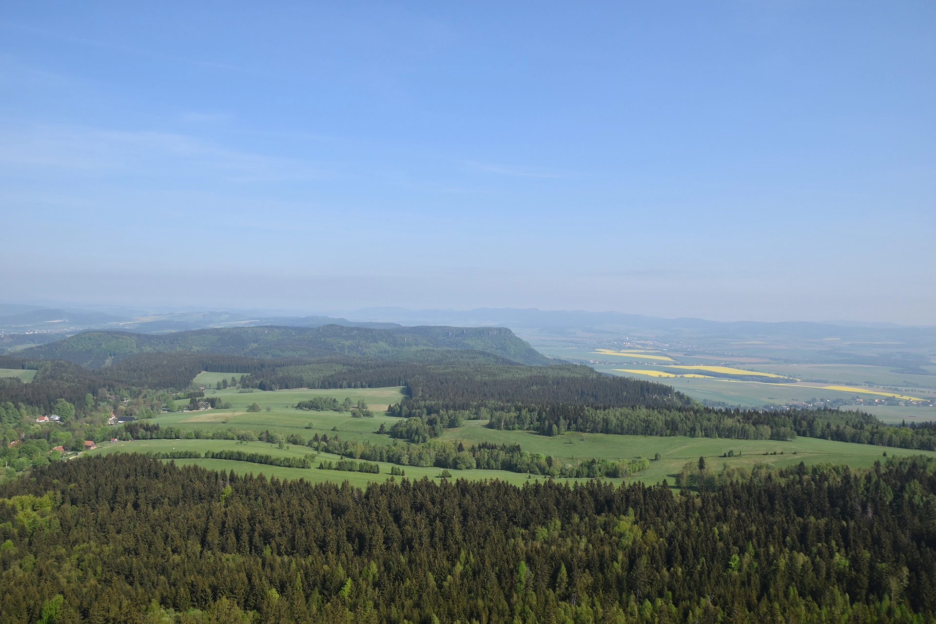 A sweeping panorama from the Table Mountains near Kudowa Zdroj reveals vibrant green fields, dense forest expanses, and distant rolling hills. The peaceful landscape stretches endlessly under a clear blue sky.