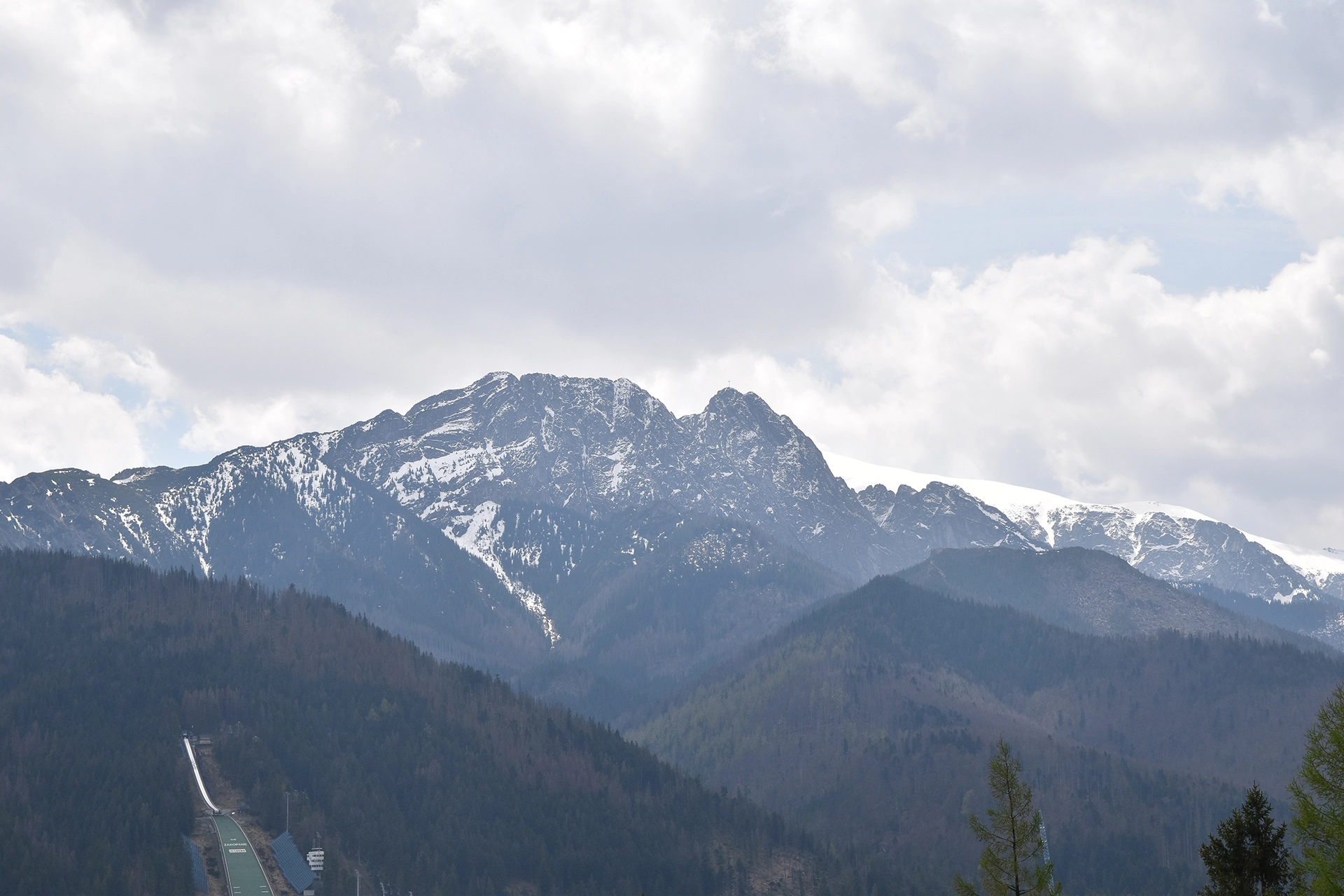 A stunning view from Gubalowka Hill in Zakopane, capturing the majestic Tatra Mountains and the iconic Wielka Krokiew ski jump nestled among the lush, forested hills.