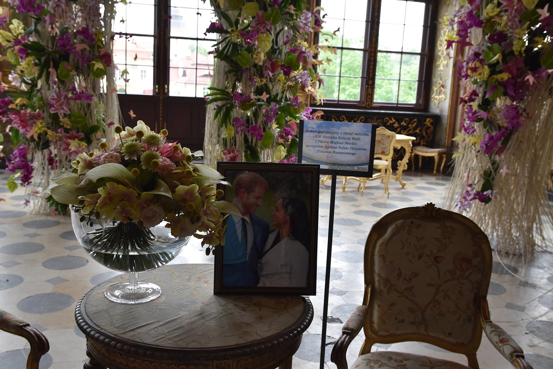 Inside Ksiaz Castle, a vibrant flower festival is underway, with stunning floral decorations adorning the elegant halls. A beautifully arranged bouquet rests on a decorative table, accompanied by a framed photo of Prince Harry and Meghan Markle. Behind the photo, a plaque offers congratulations on the occasion of their wedding, presented by Polish florists and Tomasz Kokosinski. This regal touch adds a unique, celebratory charm to the historic ambiance of the castle's interior.