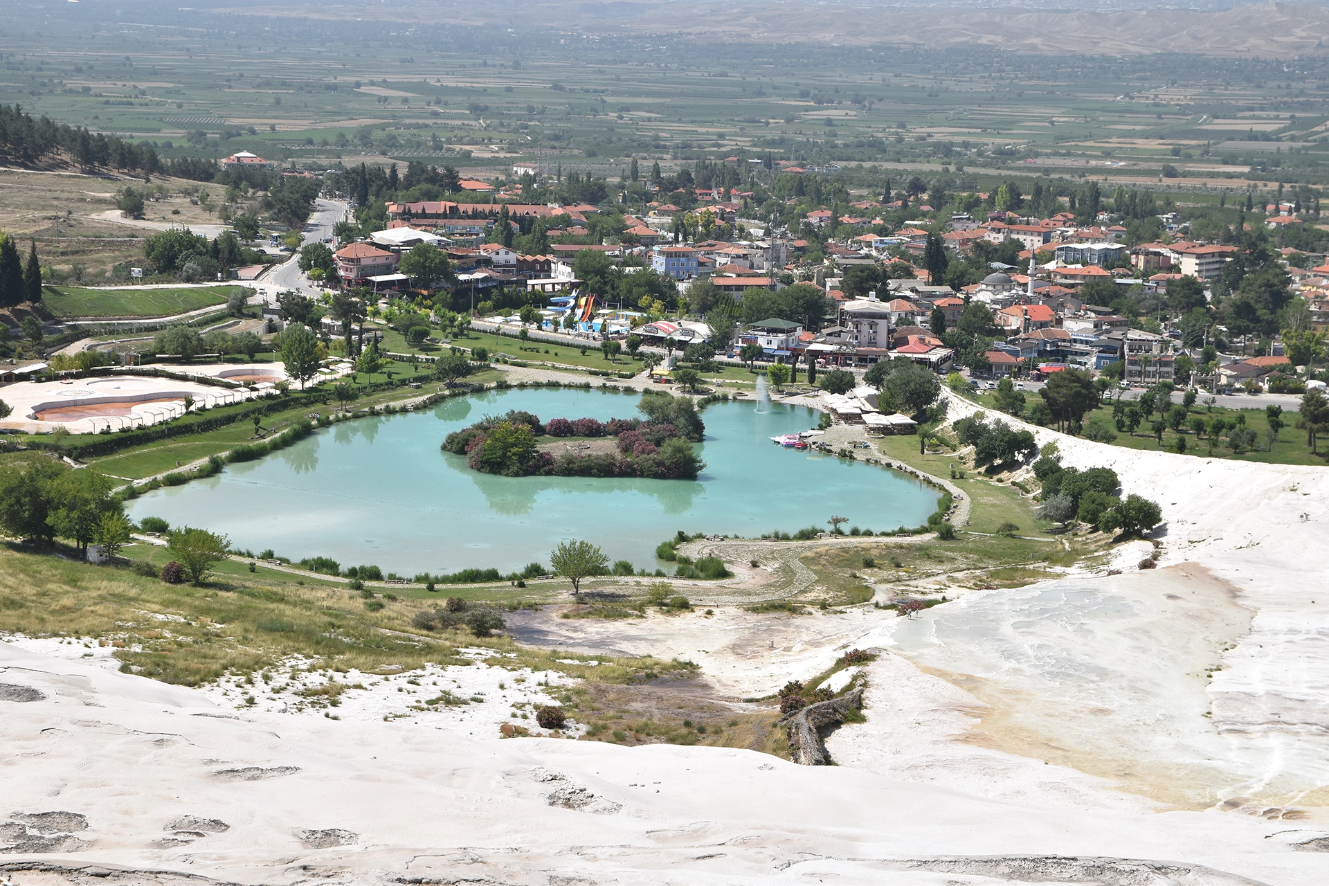 A scenic view of Pamukkale, featuring a serene lake surrounded by lush gardens and the charming village at the foot of the iconic travertine terraces.